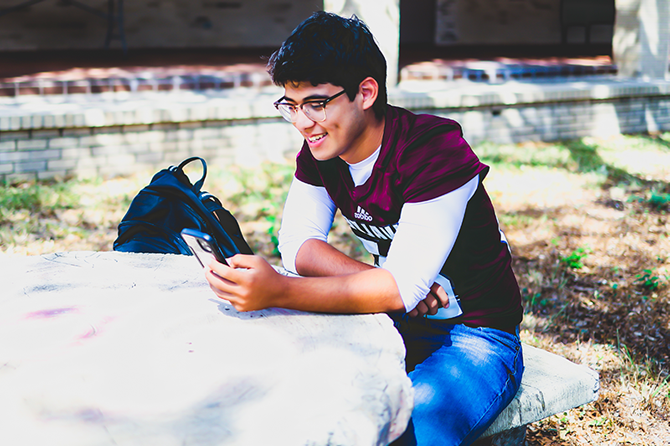 High school student sitting at a bench on campus while looking at their cell phone.